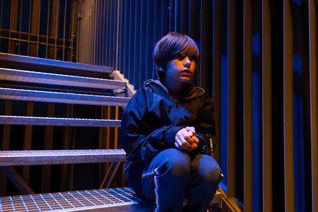 Young man sitting on stairs at night in front of a modern buildingの写真素材