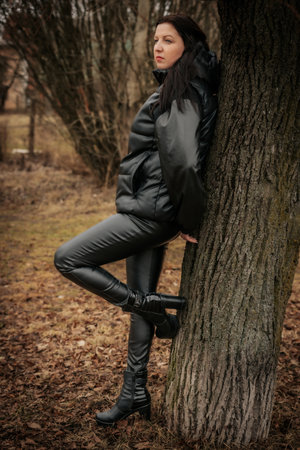Young beautiful brunette woman in a black leather jacket and boots posing near a tree.の写真素材