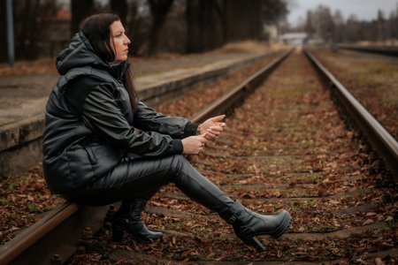 Young brunette woman in a black jacket sitting on the railway tracks.の写真素材
