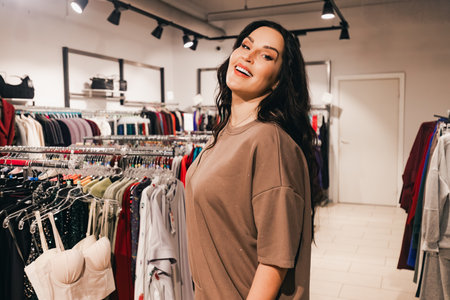 Happy woman with long dark hair in oversized brown shirt stands in clothing store, surrounded by vibrant clothing racks, embodying a joyful shopping atmosphereの写真素材
