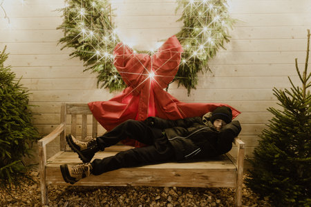 Male figure in winter clothing is relaxing on a bench adorned with festive decorations, including a large red bow and sparkling lights, evoking a warm holiday spiritの写真素材