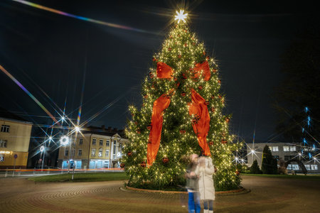 Vibrant Christmas tree illuminated with colorful lights and red bows, surrounded by blurred figures enjoying the festive ambiance in a public squareのeditorial素材
