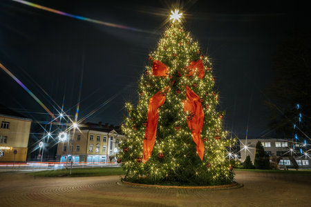 Festive Christmas tree decorated with lights and ribbons, located in a vibrant square, surrounded by holiday decorations and a beautiful night sky ambianceのeditorial素材