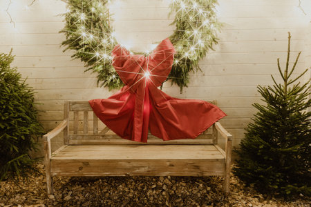 A vibrant red dress is elegantly placed on a wooden bench, surrounded by lush evergreen trees and a glowing wreath, evoking a warm holiday spiritの写真素材