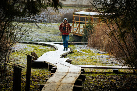 Person in red plaid jacket walks on winding wooden path through tranquil natural setting, with greenery and rustic structures, highlighting outdoor adventureの写真素材