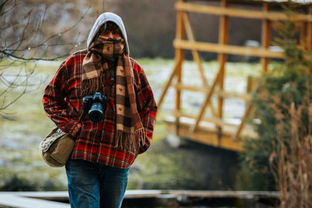 Person dressed in warm clothing, holding camera while walking along a path near wooden bridge, surrounded by greenery and natural scenery, enjoying photography outdoorsの写真素材