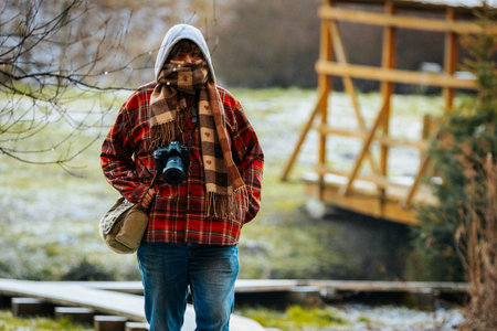 Person in cozy plaid clothing, holding camera, stands in tranquil outdoor environment, featuring wooden bridge and lush greenery, enjoying photography in natureの写真素材