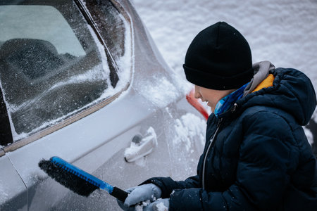 Child in winter attire is removing snow from vehicle using a brush, surrounded by a snowy landscape, demonstrating seasonal maintenance and outdoor activityの写真素材