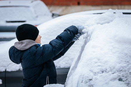 Child in winter attire is removing snow from a car windshield, surrounded by a snowy environment,...