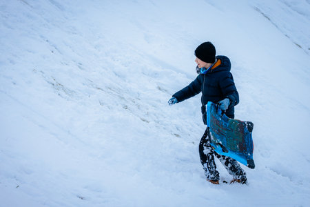 Child in winter attire climbs snowy slope with a colorful sled, showcasing joy and excitement of outdoor play in a winter wonderland filled with snowの写真素材