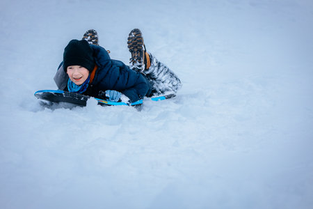 Happy young boy is sledding on a snowy hill, wearing a blue jacket and black hat, enjoying the winter season with snow-covered landscape and playful ambianceの写真素材