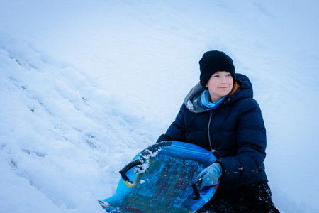 Young boy with sled sits on snowy hill, dressed in warm winter attire, smiling happily, surrounded...
