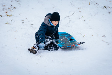 Child in winter attire sits in snow next to a vibrant sled, surrounded by a snowy landscape,...