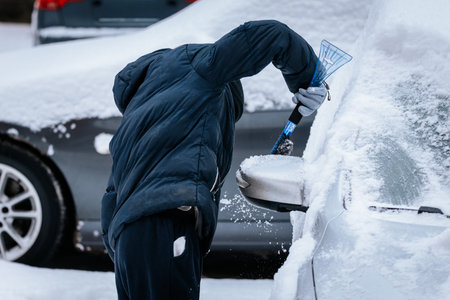 Person in winter attire is removing snow from a vehicle in a snowy setting, highlighting the effort required for winter car maintenance and outdoor activitiesの写真素材