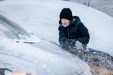Child in winter attire is actively removing snow from a vehicle hood, surrounded by a snowy...