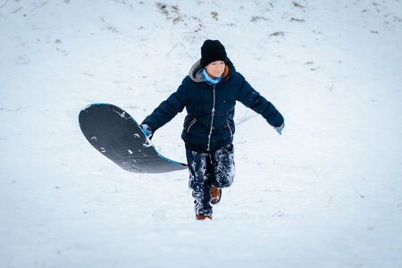 Boy in winter attire climbs snowy slope with sled, embodying excitement and adventure in winter sports, surrounded by a serene snowy landscapeの写真素材