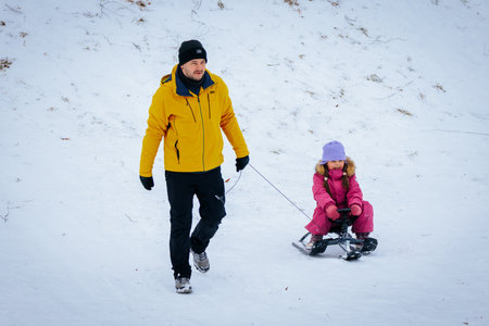 Valmiera, Latvia - January 02, 2026: Father and daughter are walking on snowy hill, child on sled,...