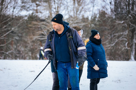 Valmiera, Latvia - January 02, 2026: Man with dog in snowy park, surrounded by friends dressed in...