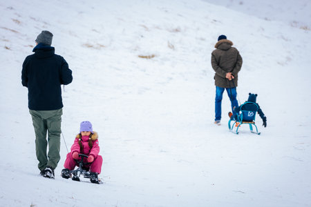 Valmiera, Latvia - January 02, 2026: Girl in pink snowsuit sleds down snowy hill while two adults...
