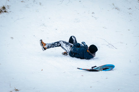 Child dressed in warm winter attire falls on snow-covered hill while sledding, surrounded by a serene winter landscape, capturing the essence of playful outdoor adventuresのeditorial素材