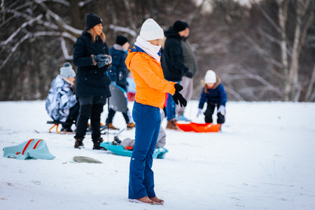 Valmiera, Latvia - January 02, 2026: Woman in orange jacket stands on snow-covered hill, preparing for sledding with family and friends in winter wonderlandのeditorial素材