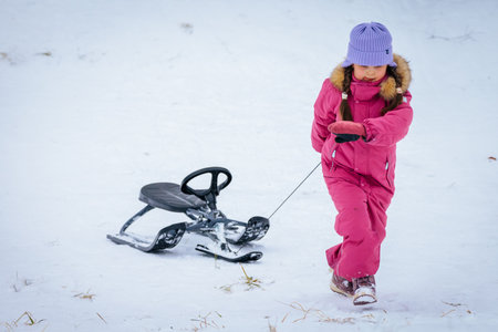 Valmiera, Latvia - January 02, 2026: Asian girl in pink coat pulls sled through snow, showcasing...