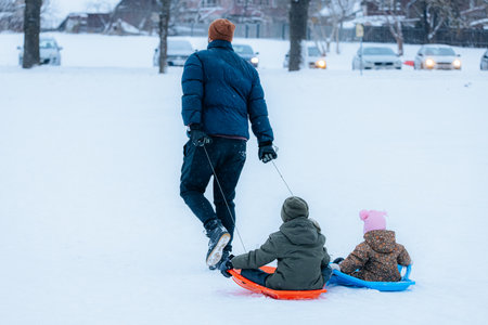 Valmiera, Latvia - January 11, 2026: Father is pulling two kids on sleds through a snowy park, surrounded by trees and a serene winter atmosphereのeditorial素材