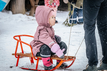 Ligatne, Latvia - January 11, 2026: Young girl in pink coat is seated on wooden sled in snow, surrounded by family enjoying winter activities and playful atmosphereのeditorial素材