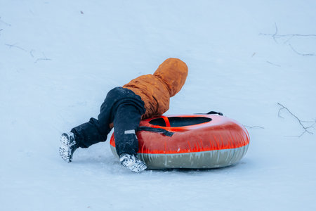 Valmiera, Latvia - January 11, 2026: Child wearing orange jacket is having fun sliding down snowy hill on inflatable tube in winter wonderlandのeditorial素材
