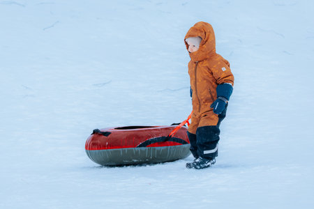 Valmiera, Latvia - January 11, 2026: Young child in orange coat is pulling red sled through snow,...