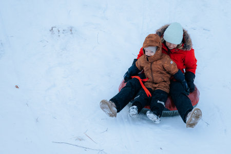 Valmiera, Latvia - January 11, 2026: Woman and young boy are sledding down a snowy slope, both wearing warm winter jackets, creating a joyful atmosphereのeditorial素材