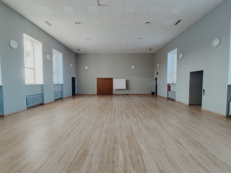 Large indoor hall features wooden flooring, large windows, and neutral walls, providing a bright and open space ideal for various events and gatheringsの写真素材