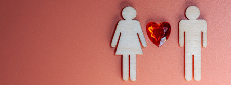 Wooden male and female figures stand beside a red heart on a coral background, symbolizing love and...
