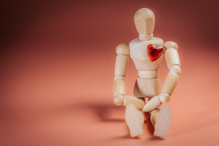Wooden mannequin figure with a red heart shape in its chest is seated against a soft orange background, representing emotions and human connection in a minimalist designの写真素材