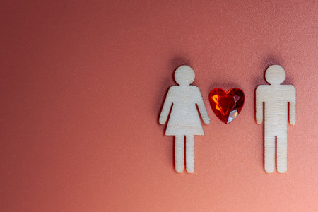 Wooden male and female figures are positioned with a red heart between them on a textured orange background, creating a visual representation of love and relationships with copy spaceの写真素材