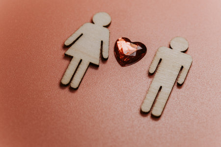 Wooden male and female figures are positioned beside a red heart-shaped gem on a soft pink surface, representing love and relationships in a creative contextの写真素材