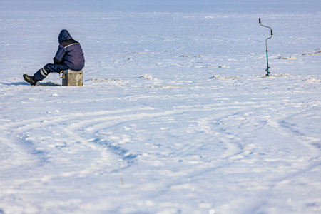 Person in winter attire sits on a wooden block on a snowy field, with ice fishing gear positioned nearby against a clear blue skyの写真素材
