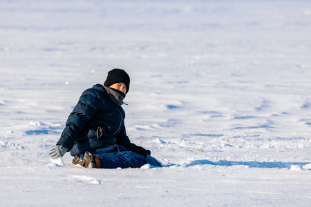 Child in winter attire sits on a snow-covered surface, smiling with a clear blue sky visible in the...