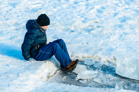 Child dressed in winter attire sits on the edge of a frozen lake, surrounded by snow and ice, with clear blue sky above and ice chunks nearbyの写真素材