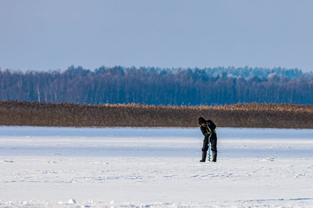 Burtnieks, Latvia - February 09, 2026: Person in black winter gear inspects the snow-covered ground in a cold, open landscape with trees visible in the distanceのeditorial素材