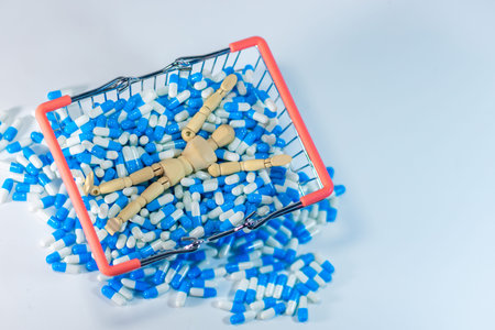 Wooden mannequin figure rests in a shopping basket surrounded by blue and white capsules, with scattered pills on a clean white backgroundの写真素材