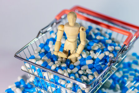 Wooden figure sits in a shopping cart overflowing with blue and white capsules, with additional pills scattered around on a light surfaceの写真素材