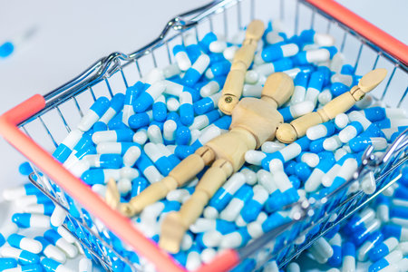 Wooden mannequin figure reclines in a shopping cart filled with blue and white capsules, with additional capsules scattered around on a white surfaceの写真素材
