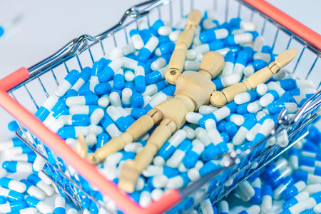 Wooden mannequin figure rests in a shopping basket filled with blue and white capsules, with additional pills scattered around on a light surfaceの写真素材