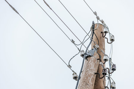 Wooden power pole supports several electrical wires and insulators against a gray sky, illustrating...