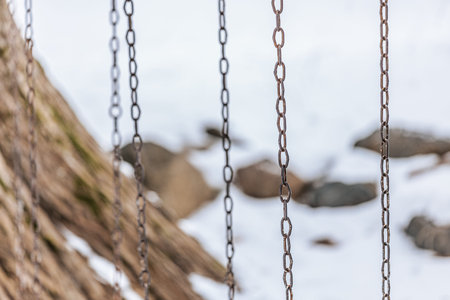 Close-up of metal chains from a swing set, with a snowy ground and rocks visible in the background,...
