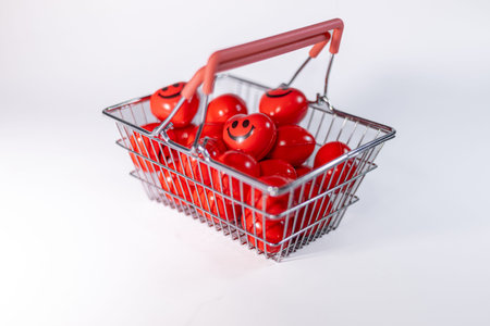Metal shopping basket contains red balls with smiley faces, set against a clean white background,...