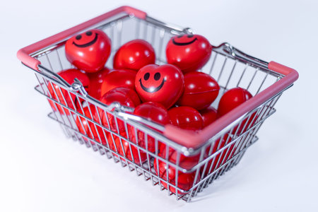 Shopping basket contains numerous red heart-shaped toys with smiling faces, set against a clean...