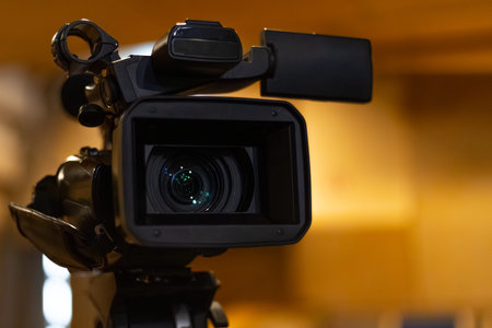 Video camera with a prominent lens is positioned on a tripod in a studio, featuring blurred...