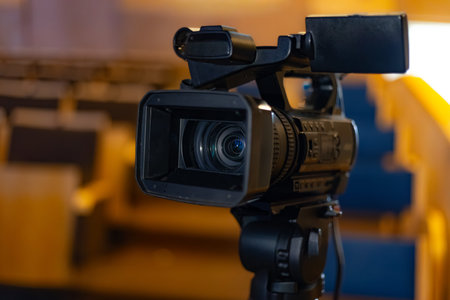 Video camera on tripod in an auditorium, featuring empty seats and warm lighting, prepared for...